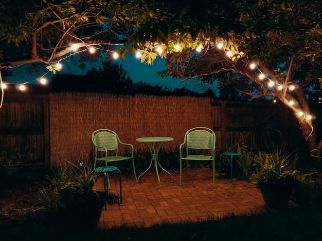 A nighttime shot of a backyard patio illuminated by a string light canopy. The lights are arranged in a crisscross pattern, creating a warm and inviting ambiance. There are comfortable chairs and a small table arranged beneath the lights, suggesting a cozy spot for relaxation and conversation.