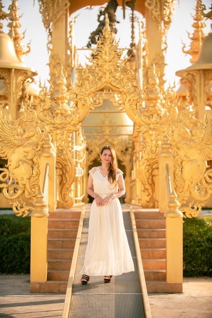 A young Thai woman, dressed in an elegant, traditional Thai dress, standing in front of a historical palace, looking determined.