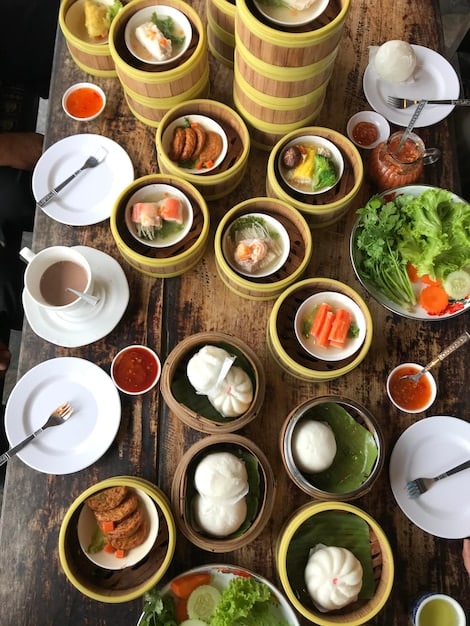 An overhead shot of a table laden with a variety of Chinese dishes, including steaming baskets of dumplings (xiaolongbao), a colorful plate of stir-fried noodles with vegetables and meat, a vibrant red bowl of mapo tofu, and several small dishes of dipping sauces and condiments. The table is set with traditional Chinese tableware and chopsticks.